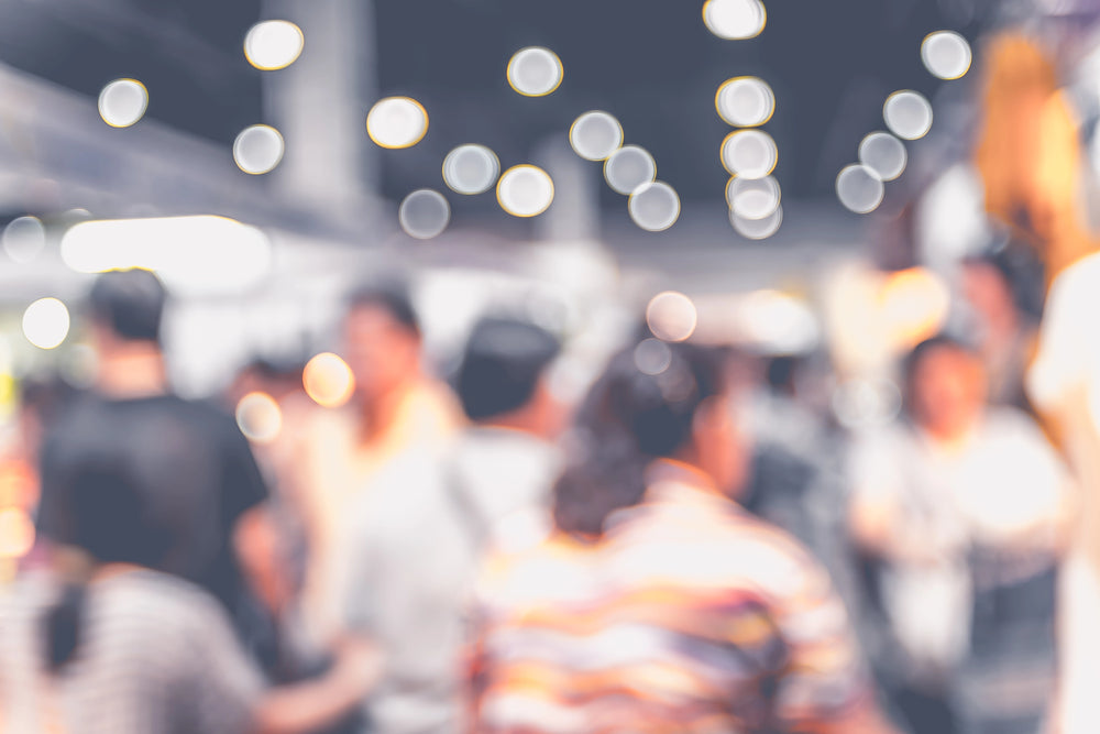Crowd moving through a busy indoor market or exhibition hall under bright overhead bokeh lights.