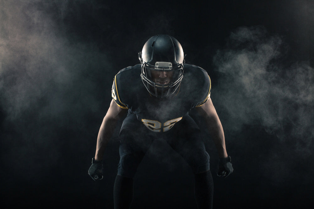 Football player crouches in a ready stance amid swirling smoke in a dim, dramatic studio setting.