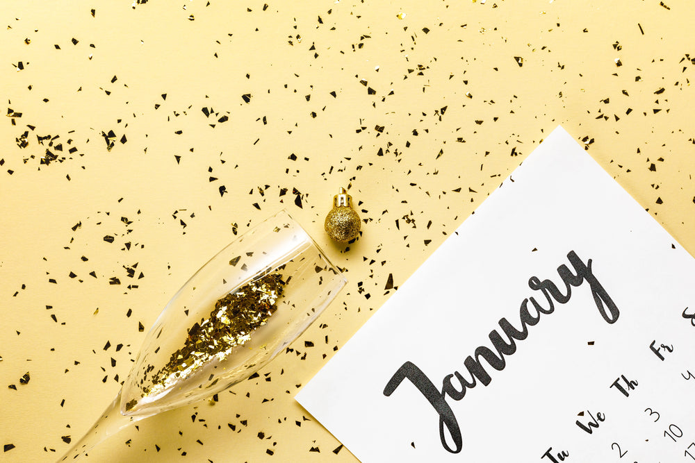 Champagne flute tipping, releasing gold confetti onto a calendar labeled "January" on a pale yellow tabletop scattered with confetti.