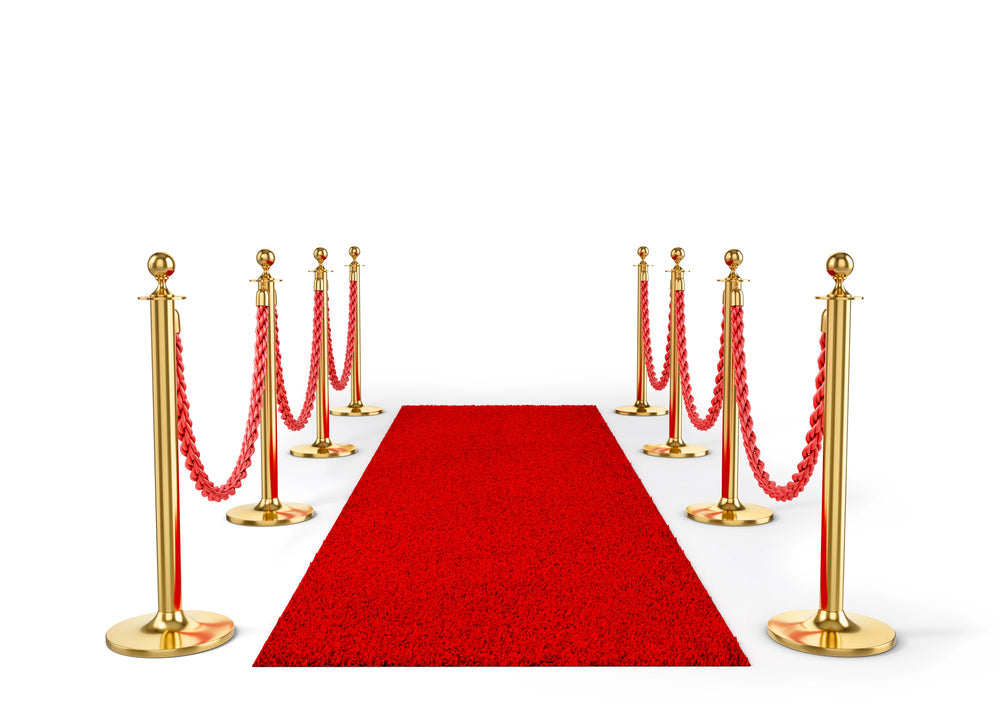 Red carpet leads between gold stanchions linked by red ropes against a white background, suggesting a formal entrance or ceremonial walkway.