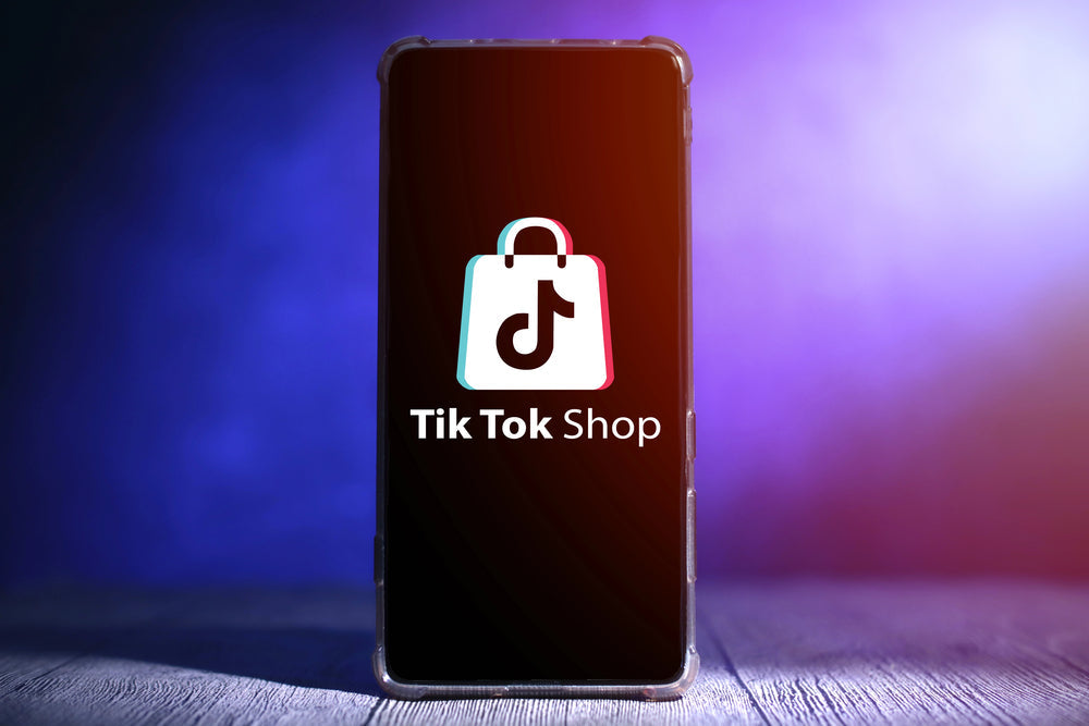 Smartphone displaying the TikTok Shop logo on its screen, standing on a wooden surface against a purple-blue gradient background.