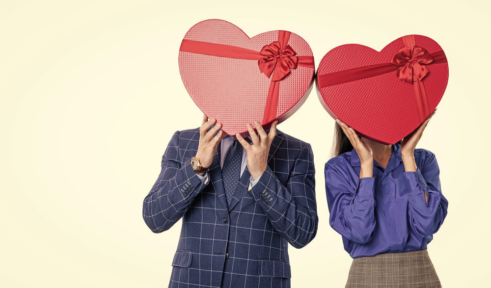 Two people hold heart-shaped gift boxes covering their faces, standing together against a light neutral background while wearing semi-formal attire.
