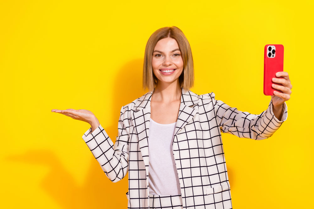 Woman takes a selfie while gesturing with her palm up against a bright yellow background.