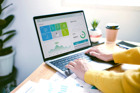 Person typing on a laptop displaying a business dashboard, working at a wooden desk with a coffee cup, potted plant, and scattered documents.