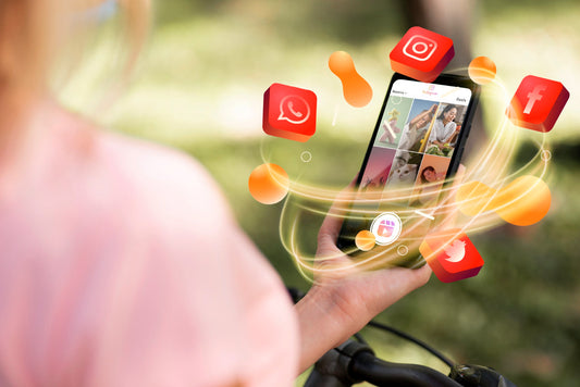 Hand holding smartphone viewing a social-media feed, app icons swirling around, outdoors with blurred greenery and bicycle handlebars visible.