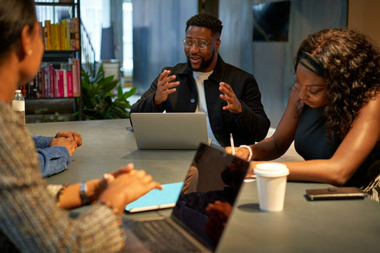 Man gestures while speaking; colleagues take notes around a conference table in a modern office with laptops and bookshelves.