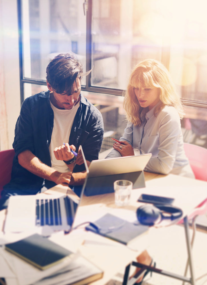 Two colleagues reviewing a tablet and laptop, pointing and discussing at a sunlit office table with notebooks and headphones by a large window.
