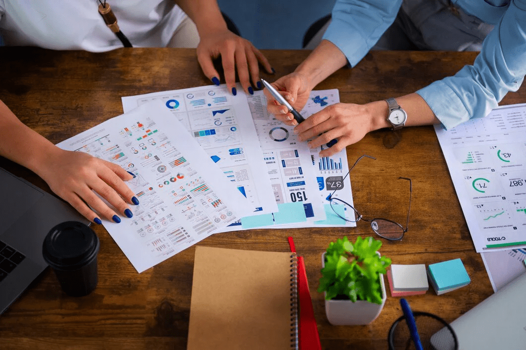 Two people reviewing printed charts and infographics, pointing and discussing data at a wooden desk surrounded by glasses, a notebook, sticky notes, a coffee cup, and a small plant.