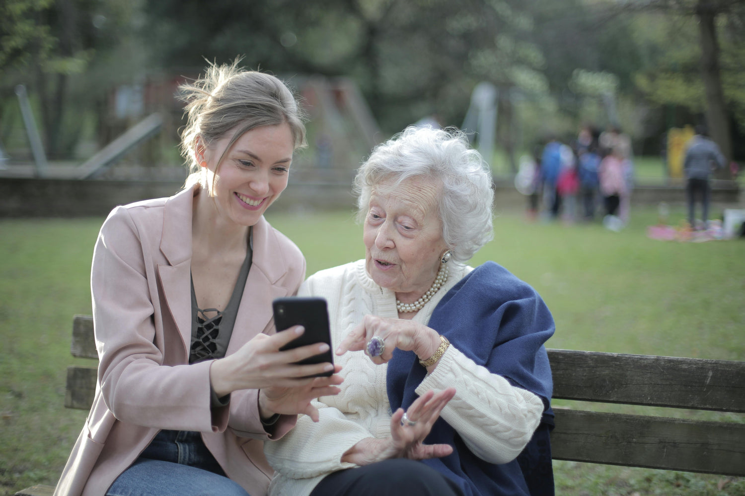 Younger woman shows a smartphone to an elderly woman while they sit together on a park bench in a playground-lined green space.