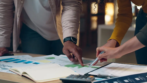 Two colleagues point and annotate charts and spreadsheets on a table during a collaborative work session in a softly lit office.